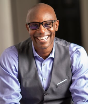 This image displays a headshot of Shola Richards, wearing glasses and a warm, welcoming smile. He is dressed in professional attire.