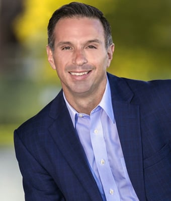Damon West in a professional headshot, wearing a dark suit and light blue shirt against a softly blurred greenery background, exuding confidence.